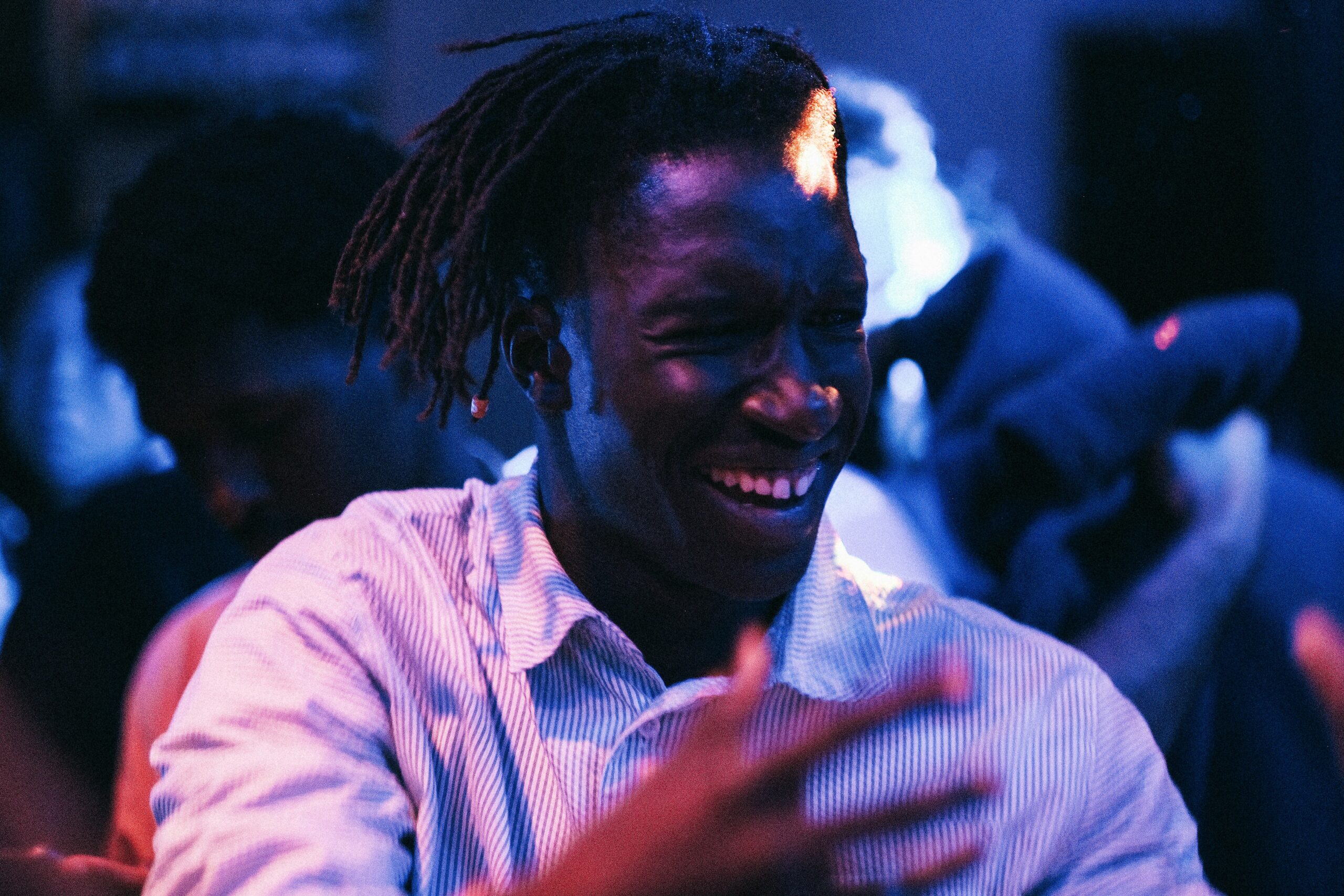 Smiling Black man with dreadlocks dancing and moving expressively in a dimly lit worship or concert setting, illuminated by blue and purple stage lights.