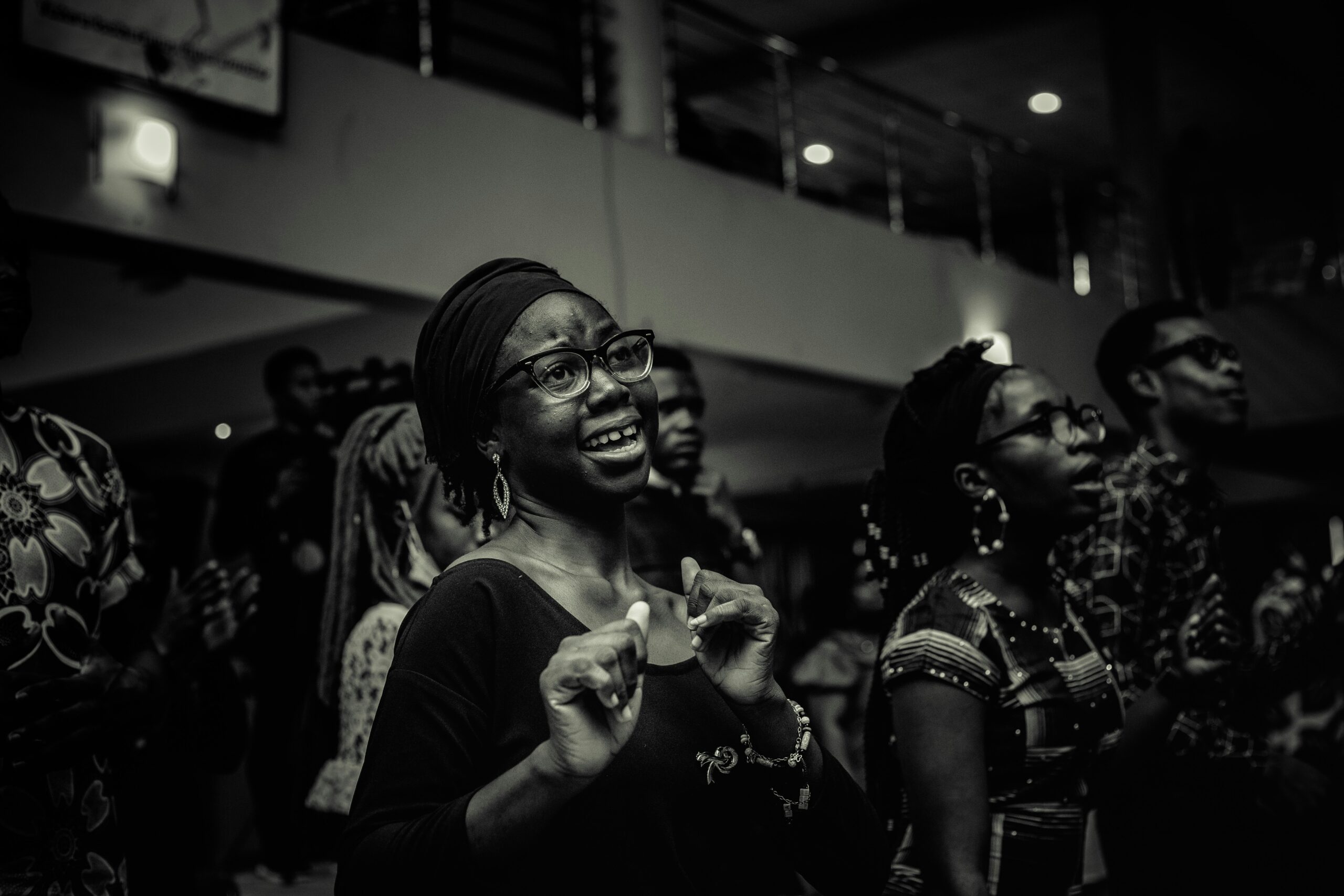 Black and white photo of a joyful Black woman wearing glasses and a headwrap, smiling and raising her hands in worship during a church service, surrounded by other believers.