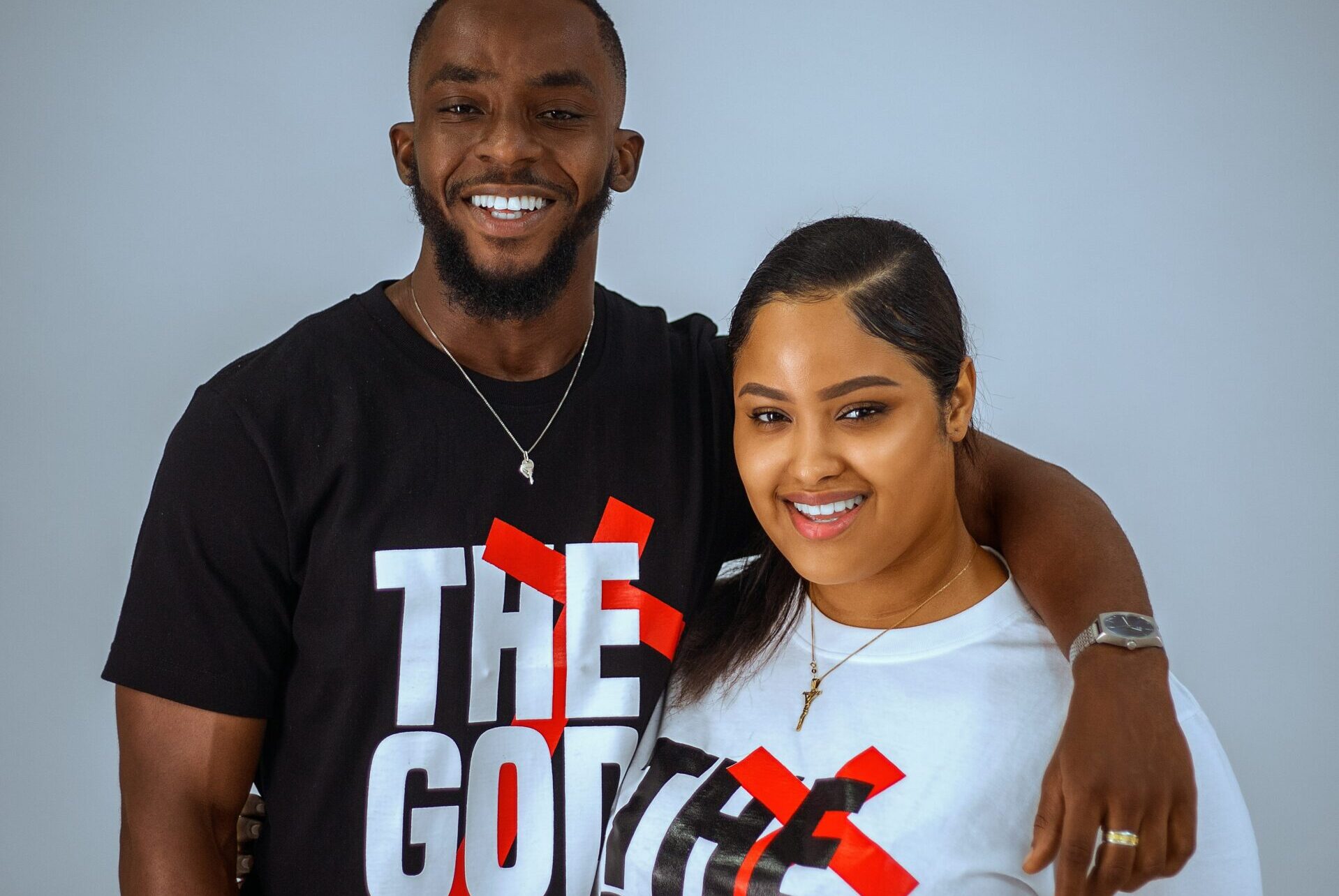 Smiling Black man and woman standing together, wearing matching 'The God Life' t-shirts in black and white, symbolizing unity, faith, and Christian lifestyle.