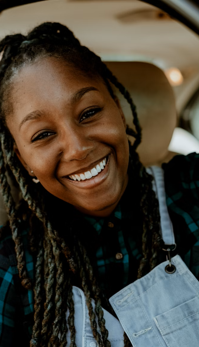 A joyful Black woman with long locs and a warm smile sits in the driver's seat of a car, wearing a plaid flannel shirt and denim overalls while looking happily at the camera.