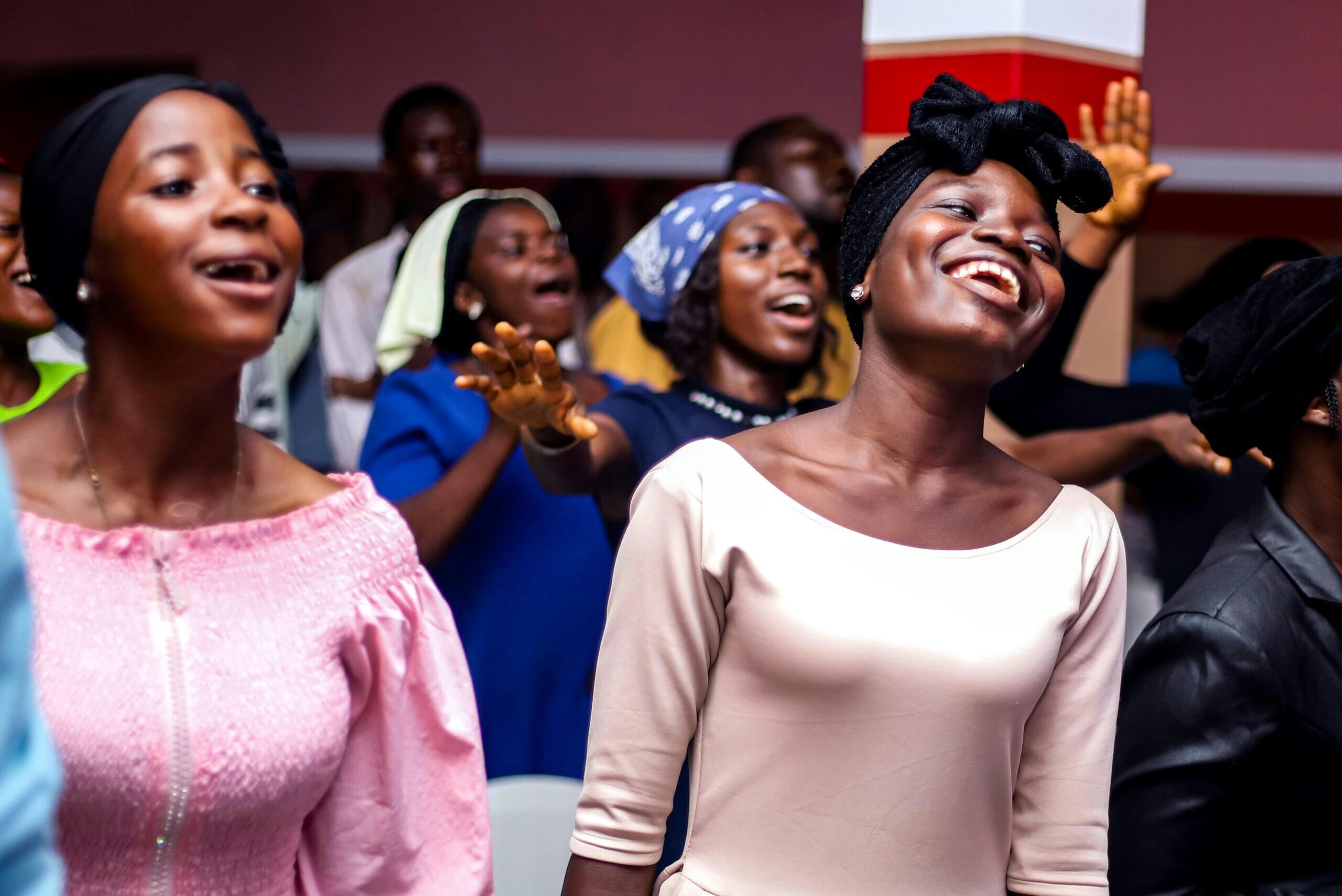 Smiling Black women with headwraps and colorful clothing worshipping joyfully in a church service, surrounded by other congregants singing and raising their hands in praise.