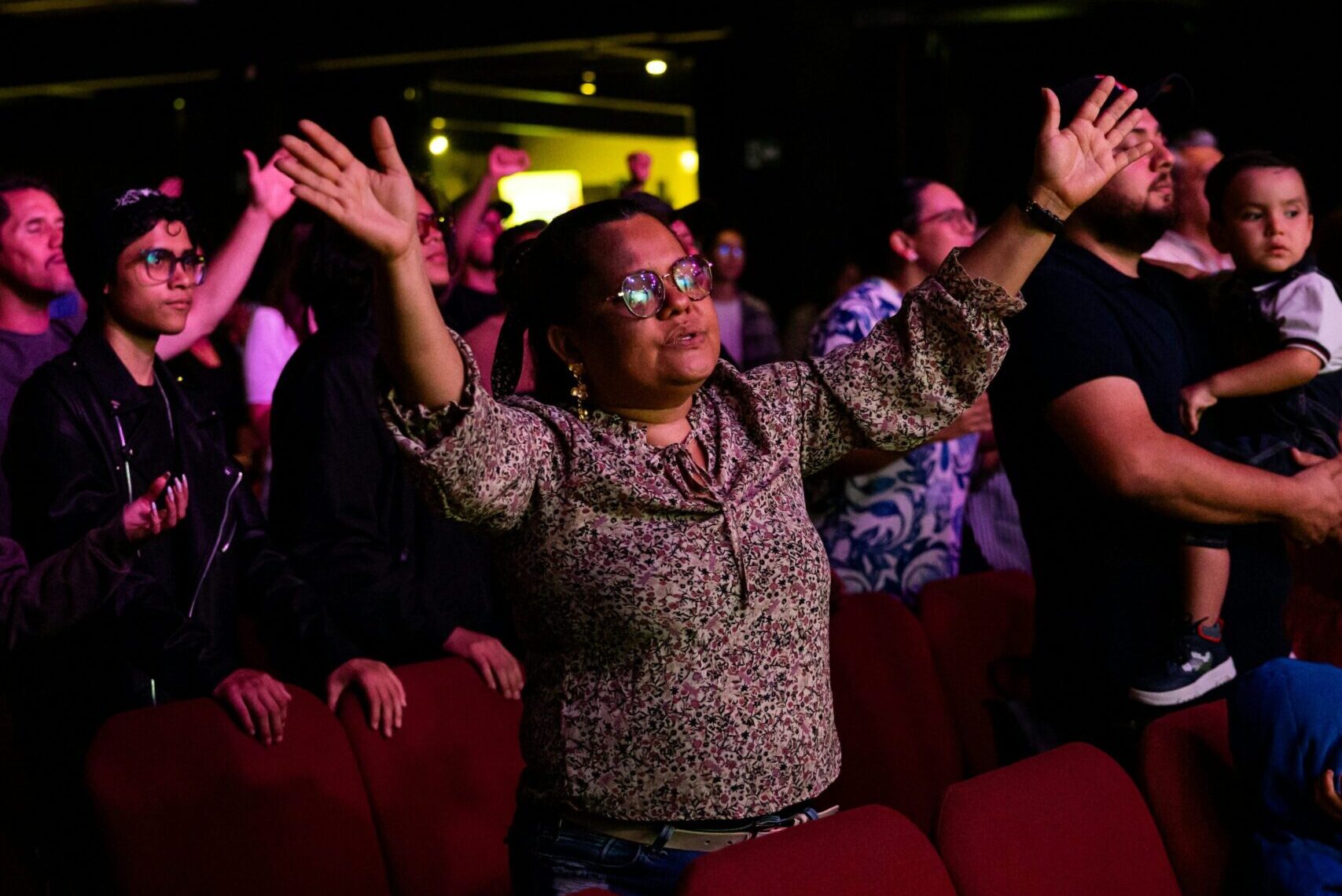 Middle-aged woman in glasses and a floral blouse standing among red auditorium chairs, raising her hands in worship during a church service, surrounded by other congregants.