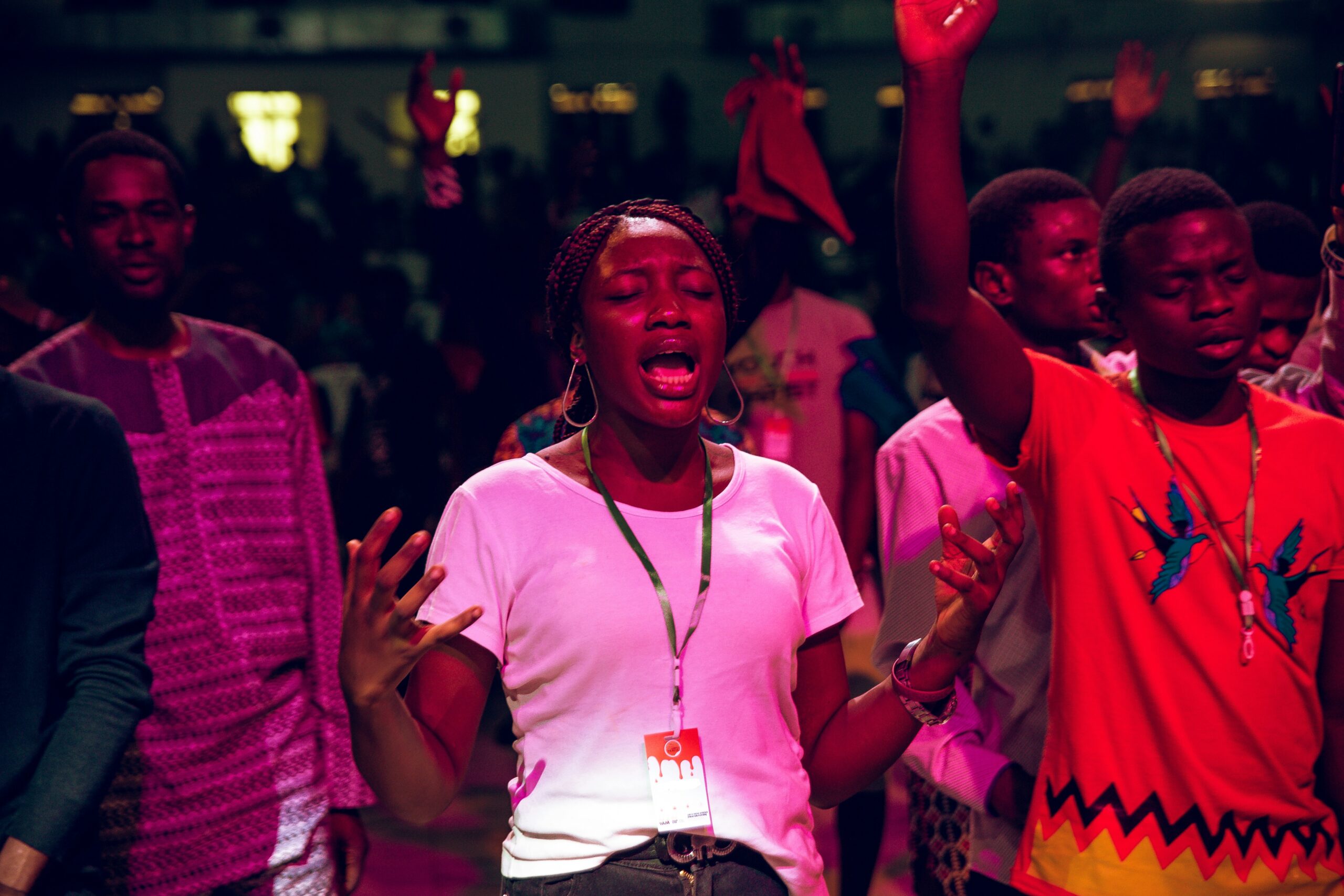 Young Black woman with braided hair and eyes closed, wearing a white t-shirt, passionately worshipping with hands raised during a vibrant church event, surrounded by other believers in colorful attire.