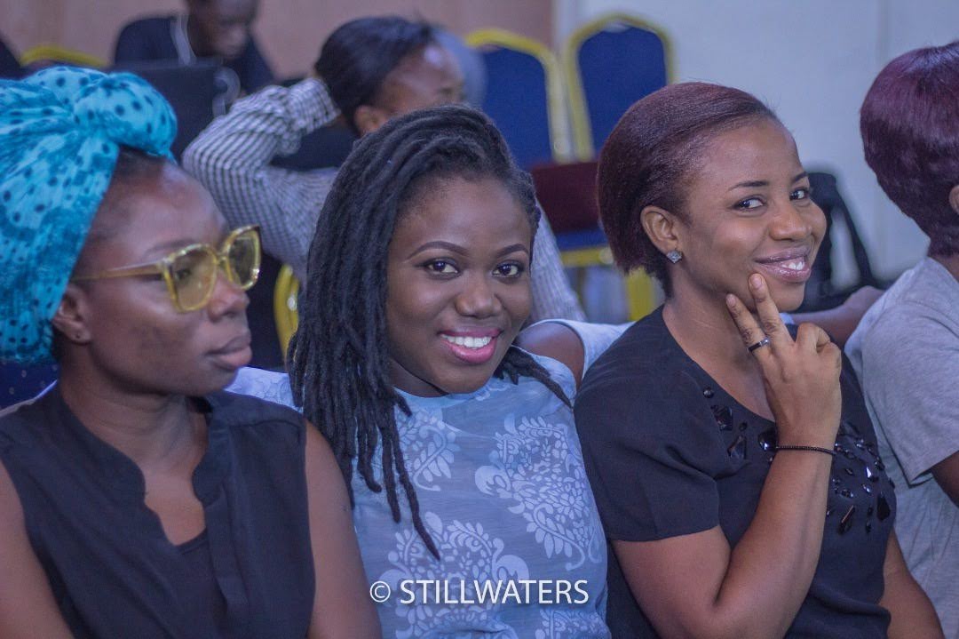 Three women smiling while seated in the congregation at a Stillwaters Ministries conference, enjoying the event.