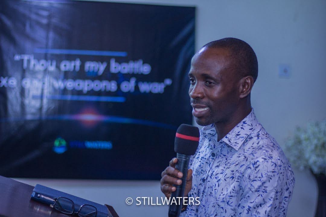A speaker delivers a message at a Stillwaters Ministries event, with a presentation screen behind him that reads, "Thou art my battle axe and weapons of war."