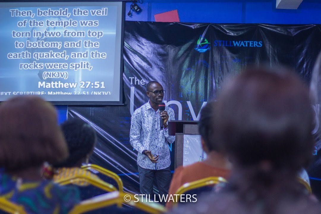A pastor teaching from a podium with a microphone at a Stillwaters Ministries event. A large screen behind him displays the Bible verse Matthew 27:51.