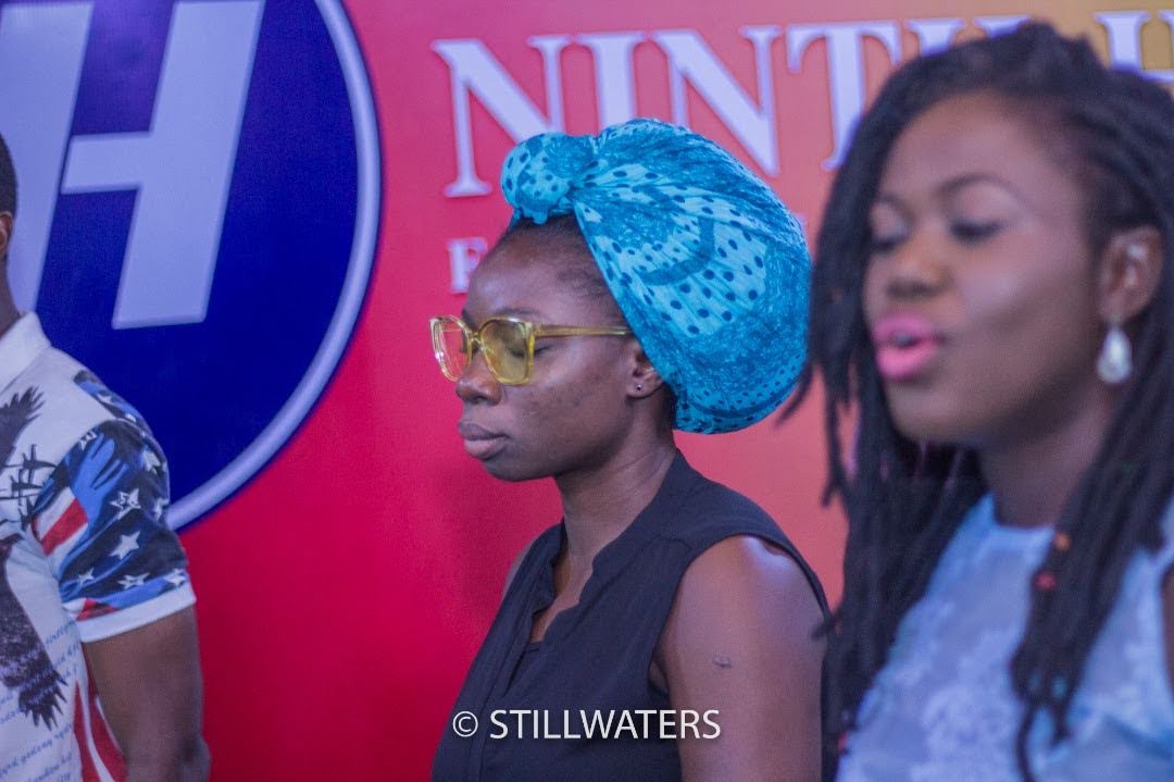 A Black woman wearing a blue headwrap and glasses in prayerful worship during a Stillwaters Ministries service, with other congregation members visible.